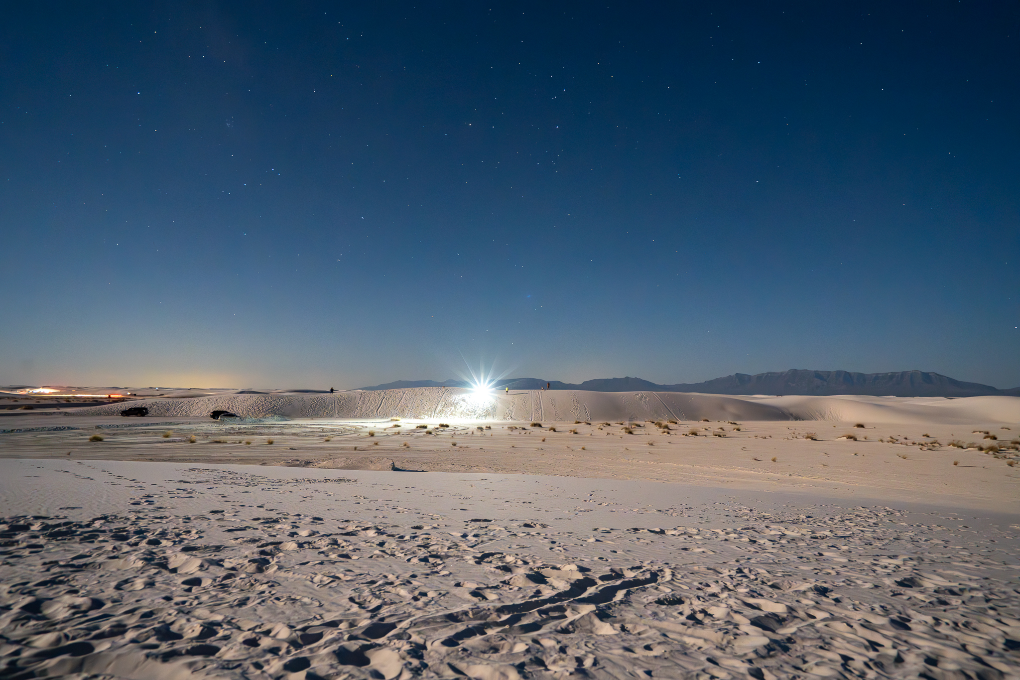 Pale sand dunes stretch beneath a deep, star-filled night sky, with a bright light glowing near a distant mountain silhouette.